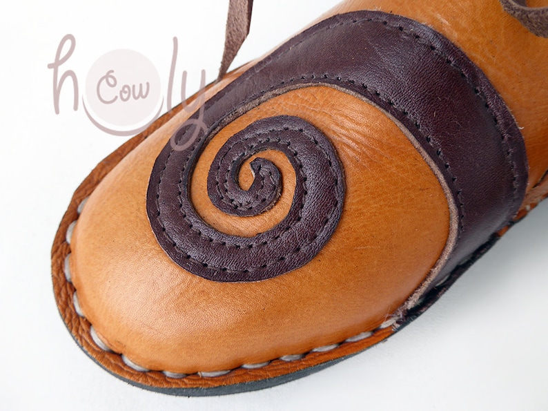 K&ouml;nnte beinhalten: Nahaufnahme eines hellbraunen Lederschuhs mit einem dunkelbraunen Spiraldesign und Akzenten. Der Schuh hat eine gen&auml;hte Sohle und einen Lederriemen. Das Design erinnert an eine Sandale oder einen Pantoffel.