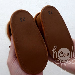 May include: A pair of brown baby shoes with a soft suede exterior. The soles are a darker brown with the number "23" printed on each. The shoes are held up by a person's hands.