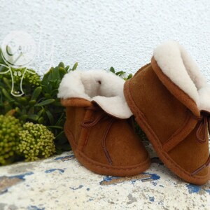 May include: A pair of brown baby booties with a soft, white, fluffy interior and trim. The booties have a suede-like exterior and are tied with brown laces. The background features green foliage and a white wall.