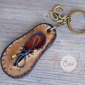 Puede incluir: Un llavero de cuero marrón en forma de sandalia con cuentas doradas y una correa de cuero negro. El llavero está unido a un anillo de metal con un dije de oro con el símbolo Om.