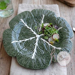 May include: A green ceramic plate in the shape of a cabbage leaf, featuring white veining. The plate is garnished with microgreens and radish slices. A fork and knife with wooden handles are on the right. A glass of water with mint is in the background.