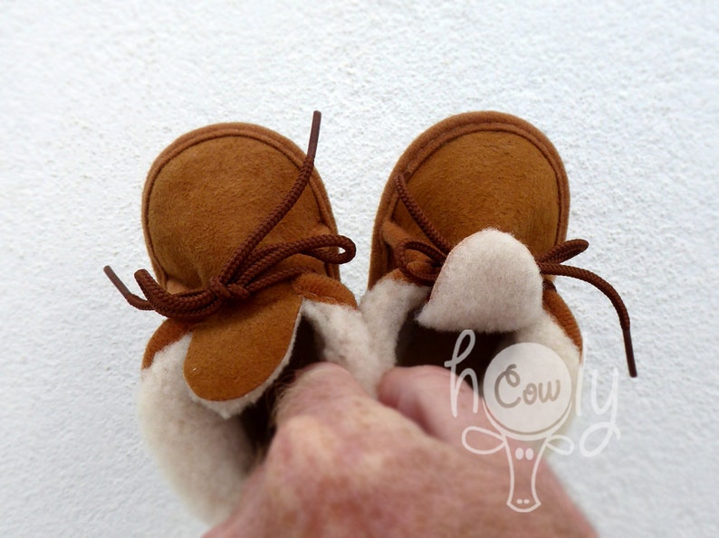 May include: A pair of brown suede baby moccasins with cream-colored fleece lining and brown laces. The shoes are designed with a classic moccasin style, perfect for infants. The image is set against a white background.