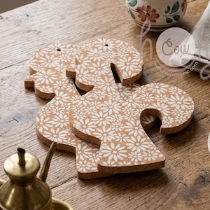 Puede incluir: Posavasos de corcho con forma original, con un estampado de margaritas blancas sobre fondo de corcho natural. Se muestran sobre una mesa de madera, ideales para el hogar o la cocina.
