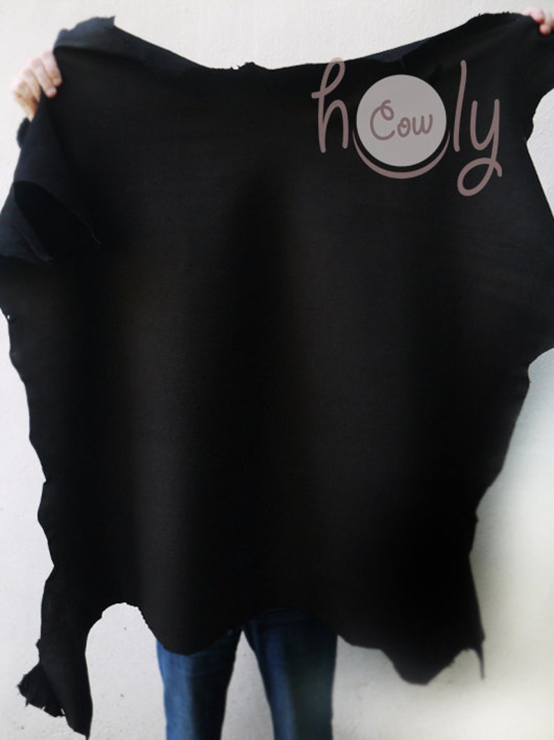 May include: A large, irregularly shaped piece of black leather is held up against a white background. The leather has a rough, unfinished edge. The text "holy cow" is in the upper right corner, in a cursive font.
