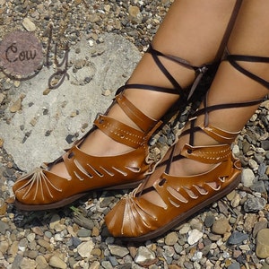May include: Brown leather sandals with laces that tie up the leg. The sandals have a cut-out design and are worn on a person's feet.