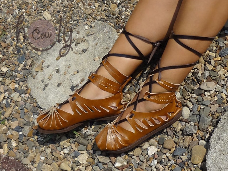 May include: Brown leather sandals with laces that tie up the leg. The sandals have a cut-out design and are worn on a person's feet.