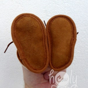 May include: A pair of brown suede baby moccasins with stitched edges and laces. The soles are a darker shade of brown. The image includes the text "h cowly".