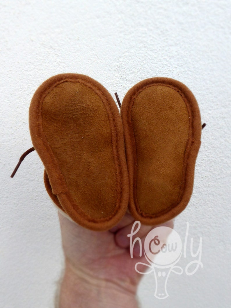 May include: A pair of brown suede baby moccasins with stitched edges and laces. The soles are a darker shade of brown. The image includes the text "h cowly".