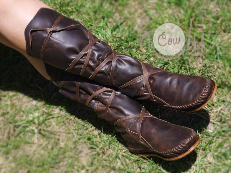 K&ouml;nnte beinhalten: Braune Lederstiefel mit Schn&uuml;rsenkeln, die sich um die Beine wickeln. Die Stiefel haben eine Mokassin-Spitze und werden auf einer grasbewachsenen Oberfl&auml;che getragen.