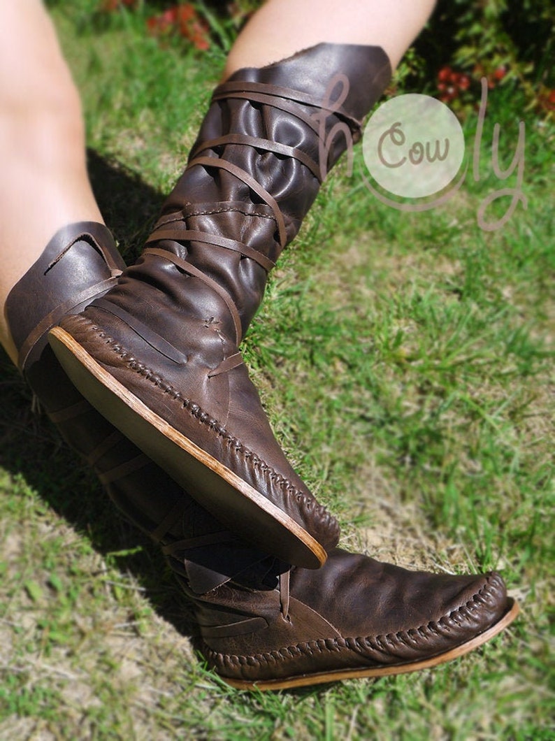 K&ouml;nnte beinhalten: Braune Lederstiefel mit Schn&uuml;rsenkeln, die um das Bein gewickelt sind. Die Stiefel haben eine Mokassin-Spitze und eine flache Sohle.