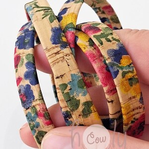 Puede incluir: Varios brazaletes de corcho con un estampado floral en azul, amarillo, rosa y verde se sostienen en una mano. Los brazaletes tienen una base de corcho natural con un diseño pintado colorido. Los brazaletes son un accesorio de moda.