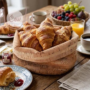 May include: A breakfast table with a basket of croissants, some with flaked almonds, on a wooden surface. Other items include coffee, juice, fruit, and jam. A plate with a croissant and jam is in the foreground.