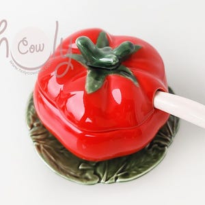 Peut inclure: Un beurrier en céramique rouge en forme de tomate avec un motif de feuille verte et une cuillère en céramique blanche. Le beurrier est posé sur une assiette en céramique verte en forme de feuille.