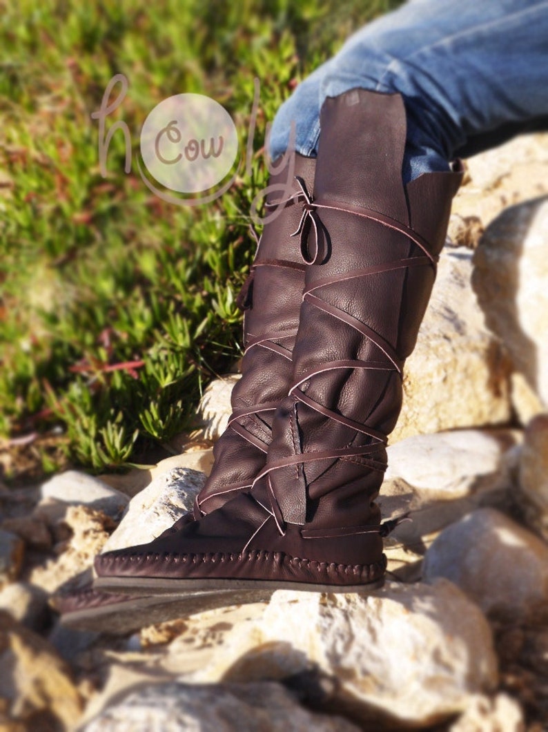 Puede incluir: Botas de cuero marr&oacute;n de ca&ntilde;a alta con cordones envueltos alrededor de las piernas. Las botas tienen una puntera de estilo mocas&iacute;n y una suela gruesa.