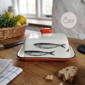 Puede incluir: Una mantequera de cerámica blanca con borde y asa rojos, con ilustraciones de peces en blanco y negro. El plato está sobre una superficie de madera, con un cuchillo y pan cerca.