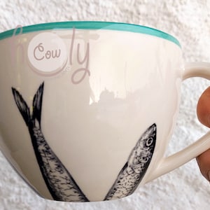 May include: Cream-colored ceramic mug with a teal rim and a curved handle. The mug features two black and white illustrations of fish. The mug is held by a hand.