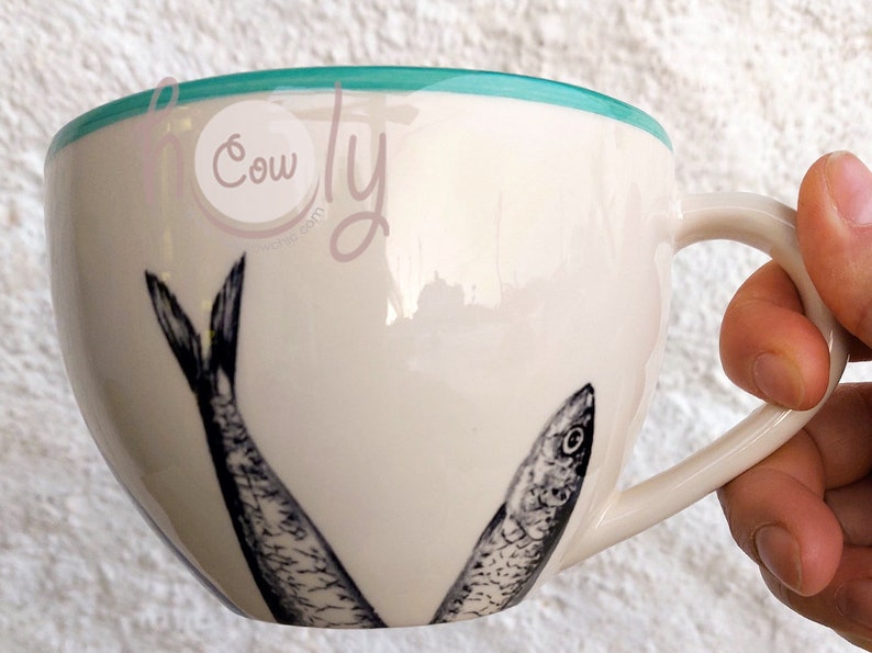 May include: Cream-colored ceramic mug with a teal rim and a curved handle. The mug features two black and white illustrations of fish. The mug is held by a hand.
