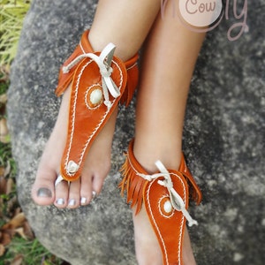 May include: A pair of brown leather sandals with fringe and white stitching. The sandals have a white leather strap that ties around the ankle. The sandals are worn on a person's feet.