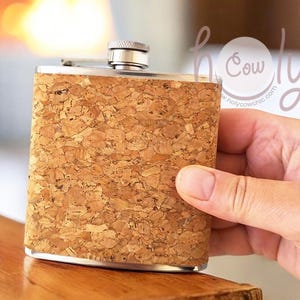 May include: A rectangular flask with a cork exterior and a silver-coloured top. The flask is held by a hand, showcasing its textured, natural cork design. The flask is designed for carrying liquids.