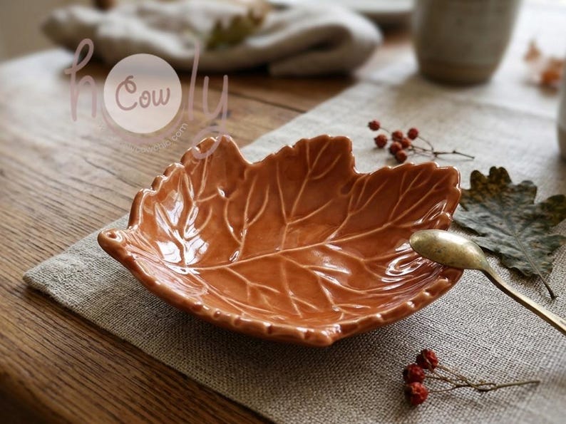 K&ouml;nnte beinhalten: Eine keramikf&ouml;rmige Schale in Form eines Blattes in einem warmen Orangebraun. Die Schale weist detaillierte Adern und ein flaches Sch&uuml;sseldesign auf. Ein goldfarbener L&ouml;ffel liegt in der Schale, daneben ein Zweig mit roten Beeren und ein gr&uuml;nes Blatt.