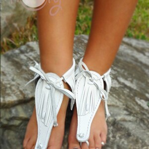 May include: White leather sandals with fringe detailing. The sandals are tied around the ankle and have a thong style toe strap.