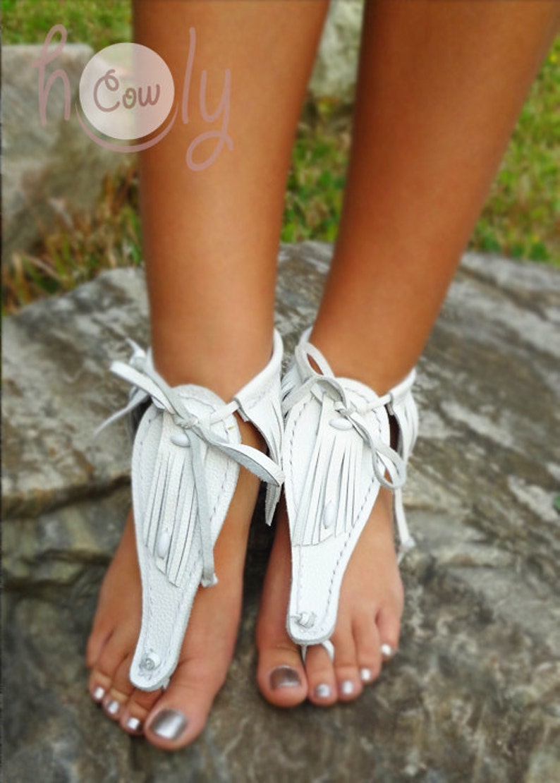 May include: White leather sandals with fringe detailing. The sandals are tied around the ankle and have a thong style toe strap.