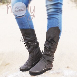 May include: Gray knee-high boots with wrap-around laces and white stitching. The boots are made of a soft material and are worn with blue jeans. The image includes the text "holy Cowly".