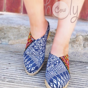 K&ouml;nnte beinhalten: Ein Paar blaue und wei&szlig;e Espadrilles mit roten und gelben Stickereien. Die Schuhe werden von einer Person getragen, die auf einer Holzoberfl&auml;che steht.