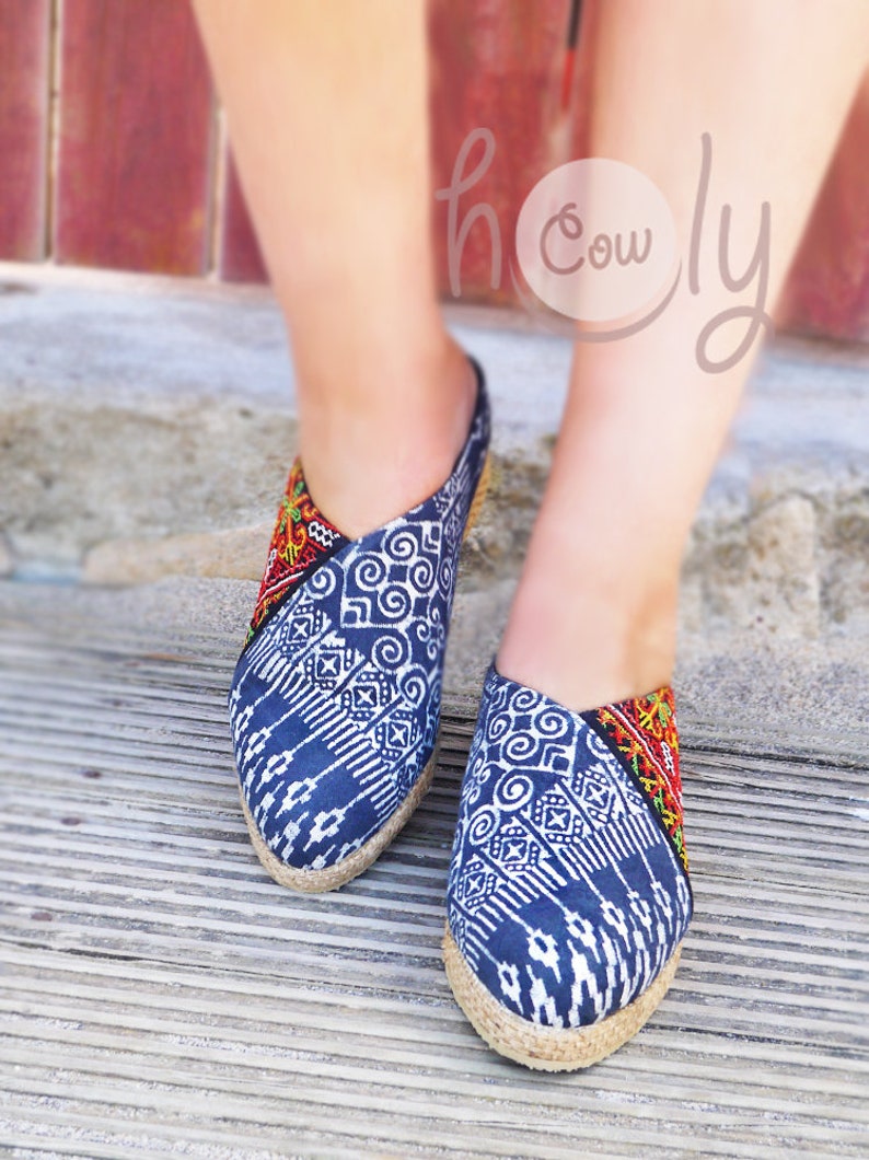 K&ouml;nnte beinhalten: Ein Paar blaue und wei&szlig;e Espadrilles mit roten und gelben Stickereien. Die Schuhe werden von einer Person getragen, die auf einer Holzoberfl&auml;che steht.