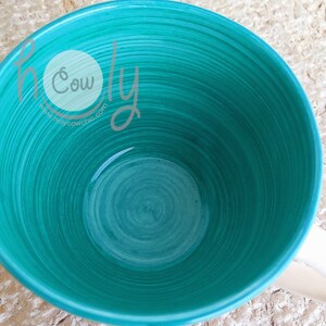 May include: A teal ceramic mug with a white handle. The interior features a spiral design in varying shades of teal. The text "holy cow chic" and a website address are visible on the side of the mug.