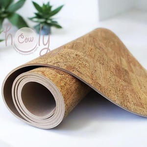 Op de afbeelding: Opgerolde yogamat van kurk. De mat is lichtbruin met een textuur. Het uitgerolde gedeelte toont een beige onderkant. Op de achtergrond groene planten en de tekst "holy cow chic".