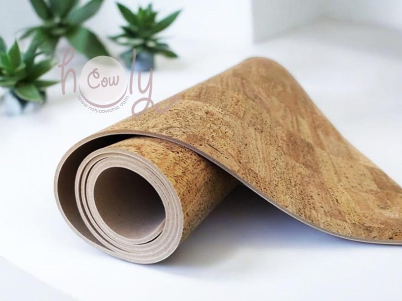 Op de afbeelding: Opgerolde yogamat van kurk. De mat is lichtbruin met een textuur. Het uitgerolde gedeelte toont een beige onderkant. Op de achtergrond groene planten en de tekst "holy cow chic".
