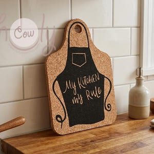 Puede incluir: Tabla de cortar de corcho con forma de delantal, con el texto "My Kitchen my Rule" en negro. La tabla está sobre una encimera de madera, con una pared de azulejos blancos al fondo.