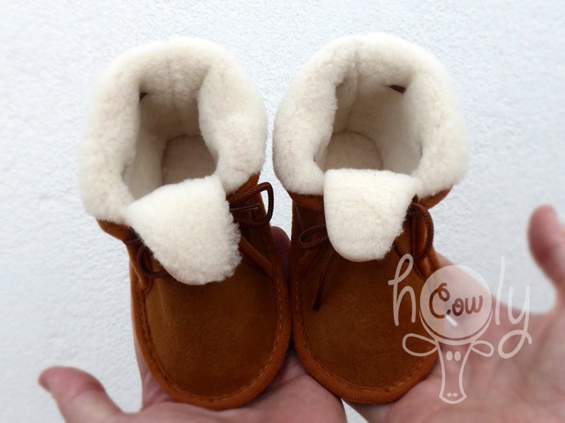 May include: A pair of brown baby booties with a soft, white, fluffy interior and trim. The booties have a leather exterior and are tied with brown laces. The overall style is a cozy, warm design.