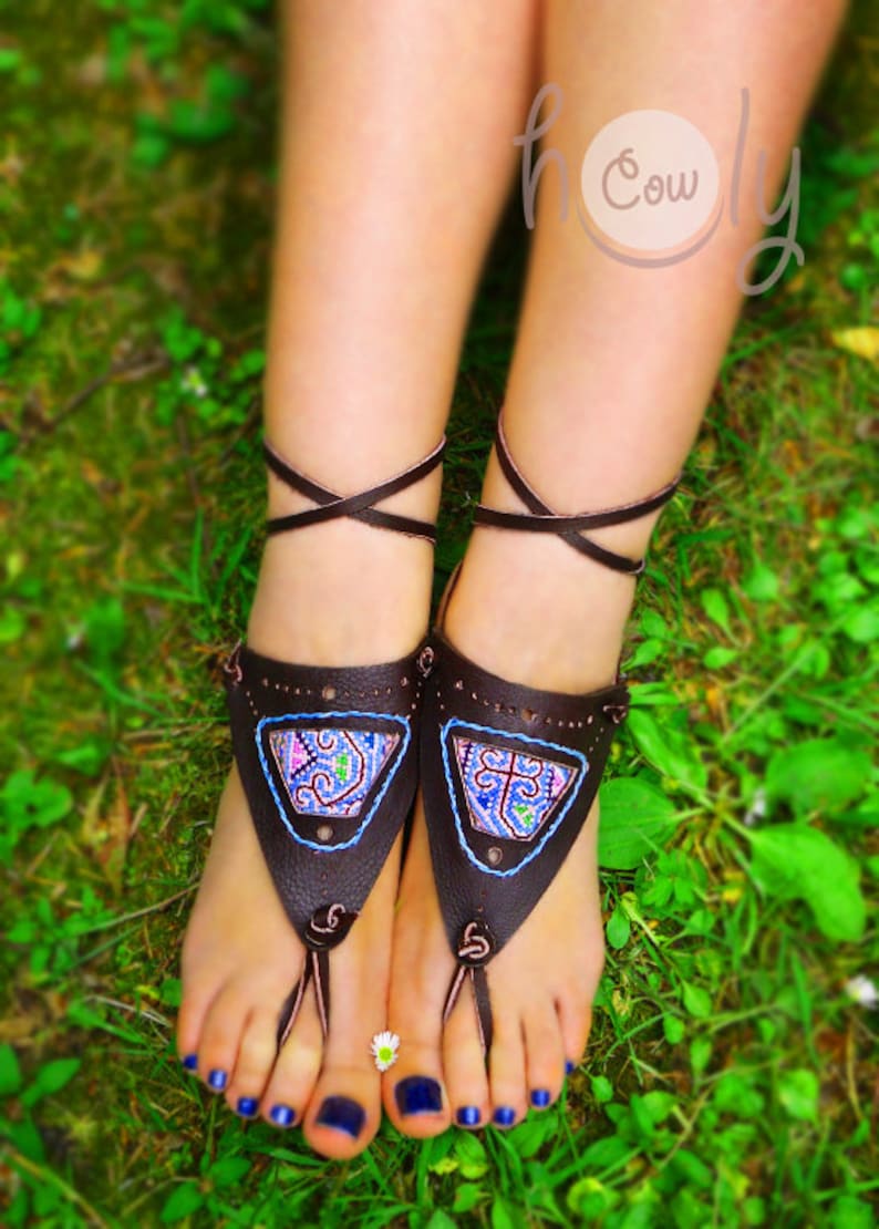May include: Brown leather sandals with intricate colorful embroidery on the front. The sandals have a toe loop and wrap around the ankle with a crisscross design. The image is taken outdoors on a bed of green grass.