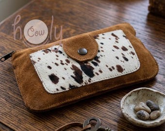 May include: A brown suede wallet with a flap closure and a section of cowhide with brown spots. The wallet has a zipper closure and a small, round button. The brand name "holy cow" is visible in the upper left corner.