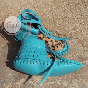 May include: A pair of turquoise leather moccasins with fringe and a leopard print insole. The moccasins are tied with leather laces.