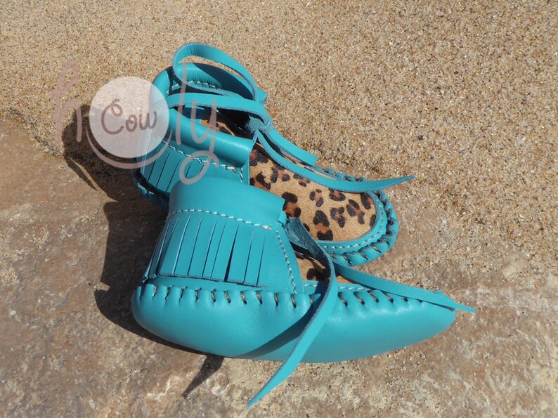 May include: A pair of turquoise leather moccasins with fringe and a leopard print insole. The moccasins are tied with leather laces.