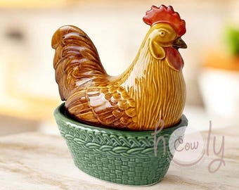 May include: A ceramic rooster figurine with a red comb and wattle, perched atop a green basket-weave base. The rooster has brown and yellow plumage. The base is a dark green color.