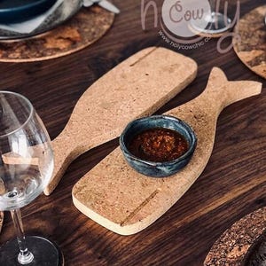 Può includere: Taglieri a forma di balena in sughero con una piccola ciotola blu piena di salsa scura. I taglieri sono su un tavolo di legno scuro, con un bicchiere da vino e un sottobicchiere visibili. I taglieri sono di colore sughero naturale.
