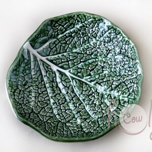 May include: A green ceramic leaf-shaped dish with a white central vein. The dish's surface is textured, replicating the intricate patterns of a leaf. This decorative item is suitable for serving food or as a display piece.