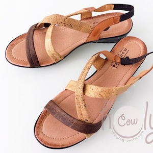 Puede incluir: Un par de sandalias de corcho y cuero marrón con una tira elástica negra. Las sandalias tienen un diseño cruzado y un tacón de cuña.