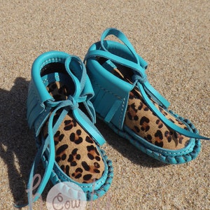 May include: A pair of turquoise blue moccasins with leopard print lining and fringe detail. The moccasins are tied with turquoise blue laces.