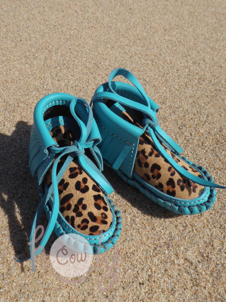 May include: A pair of turquoise blue moccasins with leopard print lining and fringe detail. The moccasins are tied with turquoise blue laces.