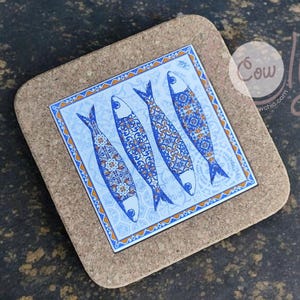 May include: Square cork coaster featuring a blue and white tile design with four stylized fish. The fish have intricate patterns and are set against a light blue background. The coaster has a natural cork border.