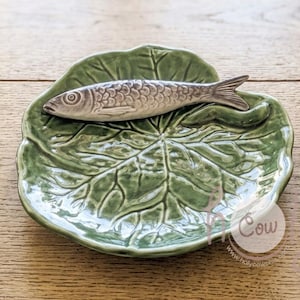 Pode incluir: Um prato de cerâmica verde em forma de folha de couve com uma figura de peixe cinzenta e texturizada em relevo no topo. O prato apresenta um acabamento brilhante e nervuras de folha detalhadas. Um logótipo circular com o texto Holy Cow é visível no canto inferior direito.