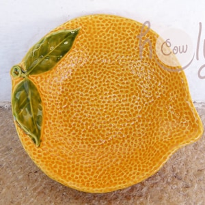 Può includere: Un piatto di ceramica giallo a forma di limone con foglie verdi. Il piatto ha una superficie testurizzata che ricorda la buccia di un limone.