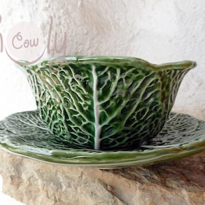 May include: A green ceramic bowl and saucer set, shaped like a cabbage. The bowl has a ruffled edge, and both pieces are textured to resemble cabbage leaves. The set is displayed on a stone surface.