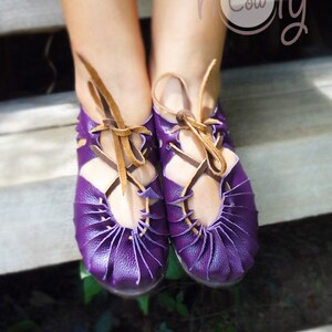 May include: Purple leather sandals with brown laces. The sandals have a unique design with multiple layers of leather. The sandals are worn by a person on a wooden surface.