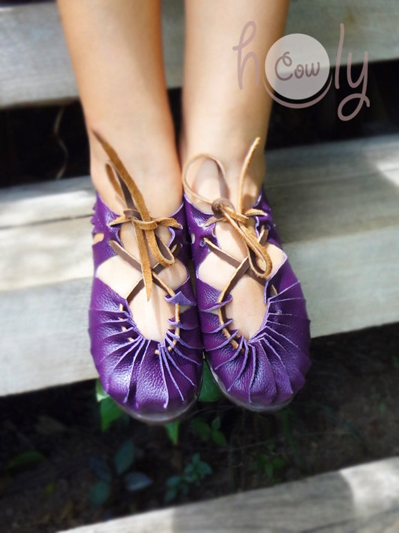 May include: Purple leather sandals with brown laces. The sandals have a unique design with multiple layers of leather. The sandals are worn by a person on a wooden surface.